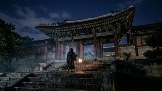 A lone figure with a paper lantern approaches a Joseon-era palace gate at night